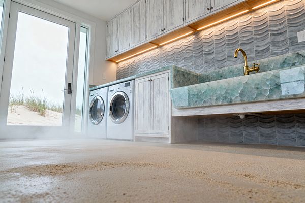 A bright coastal laundry room with a full-height glass door looking out to sand dunes features a side-by-side front-load washer and dryer beneath whitewashed wood cabinets. A large sea-glass green textured glass utility sink with a brass faucet sits against a gray-blue wave-pattern ceramic tile backsplash, highlighted by warm under-cabinet lighting, with windblown sand visible across the light floor.