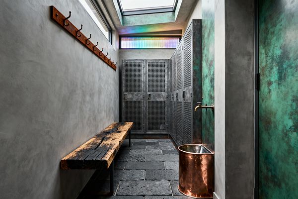 A narrow industrial mudroom with gray plaster walls and a black slate floor features a long rusted hook rail and a rough reclaimed-wood bench on the left. At the end are perforated steel lockers. On the right, verdigris copper panels frame a brass spout above a cylindrical copper basin. A skylight and a high ribbed-glass window bring daylight, casting soft shadows and a faint rainbow glow above the lockers.
