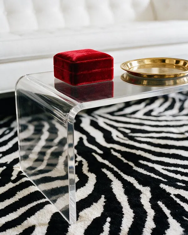 A clear acrylic side table with rounded edges sits on a black-and-white zebra rug, topped with a small red velvet box and a gold tray. A white sofa blurs softly in the background, emphasizing the glamorous, high-contrast styling.