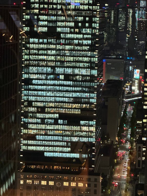 A nighttime view of a tall office building in Tokyo, showcasing illuminated windows and a bustling city street below.