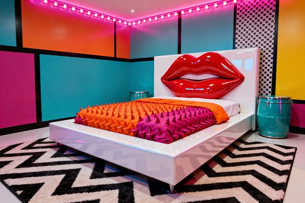 Pop art bedroom with a glossy white platform bed and an oversized red lips headboard. The bedding includes an orange duvet and a magenta throw covered in cone-shaped spikes. Walls are color-blocked in teal, tangerine, magenta, and yellow, outlined with black grid lines, with a black-on-white polka-dot panel. Pink perimeter bulbs cast neon light from the ceiling. A black-and-white chevron rug sits beside a turquoise drum nightstand and a small chrome side table.