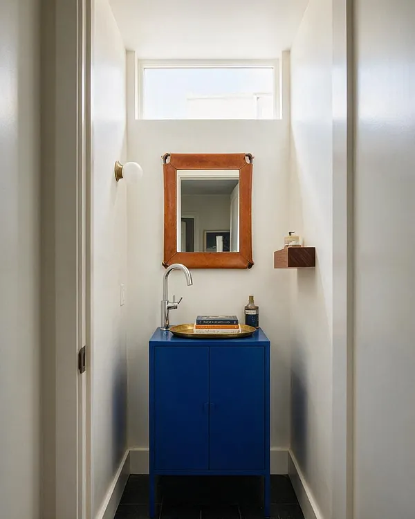 A narrow powder room with white walls, a small horizontal window near the ceiling, and a bright cobalt blue vanity centered below a copper-framed mirror. A chrome faucet rises from the sink, while a brass wall sconce, a small wooden shelf, and a black tile floor add contrast and character.
