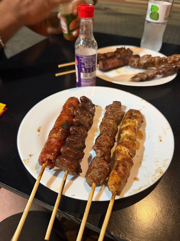 A man and a woman are sitting outdoors at a wooden table with meat skewers and glasses of beer, smiling and giving thumbs up, with a tropical outdoor setting in the background.