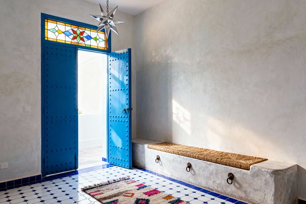 A bright mudroom features cobalt-blue, studded double doors beneath a multicolored stained-glass transom. Limewashed plaster walls frame a built-in masonry bench topped with a woven palm cushion, its front detailed with wrought-iron rings. The floor is white ceramic octagon-and-dot tile with a deep blue border and a small flatweave rug in red, pink, and navy. Daylight streams through the open door and a metal Moravian star pendant hangs above.