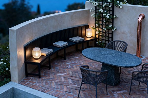 A transitional patio at dusk features a curved beige stucco wall with a black slatted bench, three rough stone seat pads, and two glowing ribbed-glass lanterns. The floor is herringbone terracotta brick. An oval dark-stone pedestal table is paired with charcoal rope-woven chairs. A black trellis supports a climbing vine with white flowers, and a copper downspout accents the corner, with distant water softly visible beyond.