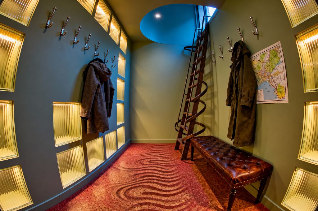 A narrow mudroom with sage green walls features a grid of backlit, fluted glass blocks that appear warped. Along both sides, stainless coat hooks hold dark coats above a tufted brown leather bench and a pinned map. At the end, a dark-wood ladder wrapped by a spiral metal band rises to a circular, sky-blue oculus skylight. Terracotta cork flooring with wavy maroon patterns amplifies the dreamlike feel while still offering practical seating and hanging space.