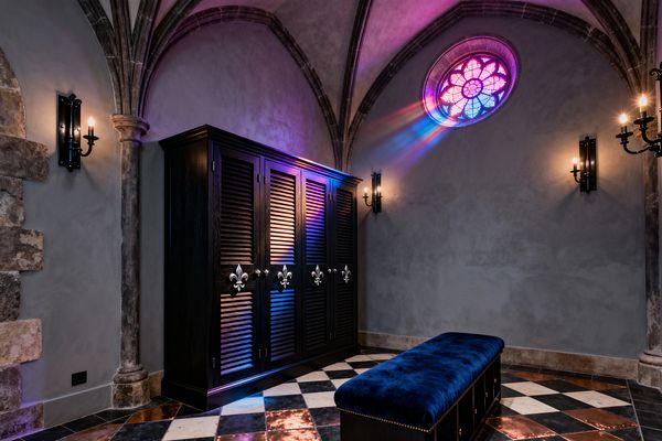 A Gothic-inspired mudroom with ribbed stone vaults and gray plaster walls features a round rose stained-glass window casting pink and blue light. A dark-stained louvered locker cabinet with fleur-de-lis pulls stands beside wrought-iron candle sconces. A deep blue velvet bench with brass nailheads rests on a black-and-white checkerboard floor dotted with copper tiles and framed by aged stone columns.