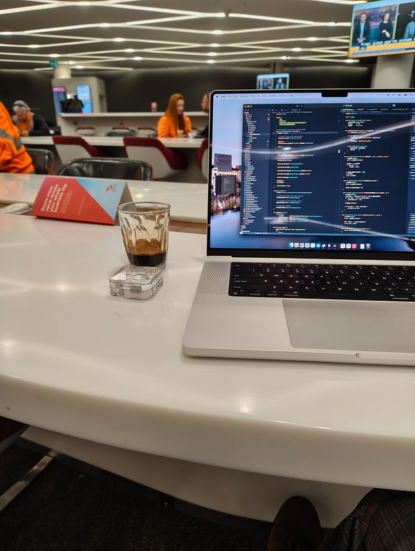 A workspace in a lounge filled with people, featuring a laptop and a coffee.
