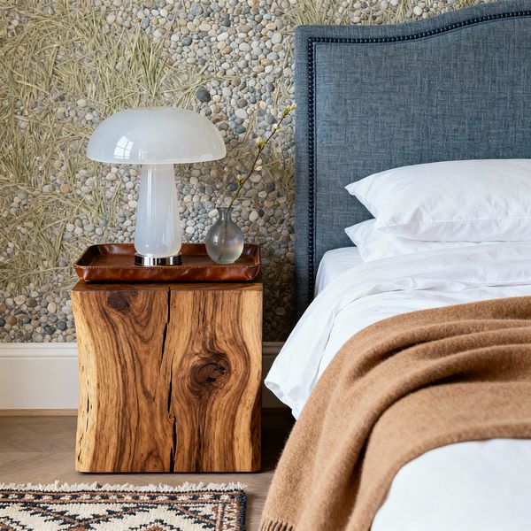 Close-up of a transitional bedroom featuring a slate-blue upholstered headboard with dark nailhead trim beside a solid live-edge wood nightstand. On the nightstand are a brown leather tray, a white opal glass mushroom lamp with a chrome base, and a small frosted glass bud vase. The wall shows pebble-and-grass photographic wallpaper. The bed has crisp white sheets with a camel wool throw, with light herringbone flooring and a fringed patterned rug visible.