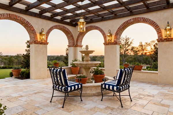 A traditional patio with a dark wood pergola spans stucco columns and red brick arches, centered on a tiered stone fountain set in a circular basin. Two wrought-iron chairs with navy-and-white striped cushions sit on travertine pavers, while brass lanterns and terracotta planters accent the space, opening to landscaped lawns and distant trees at sunset.