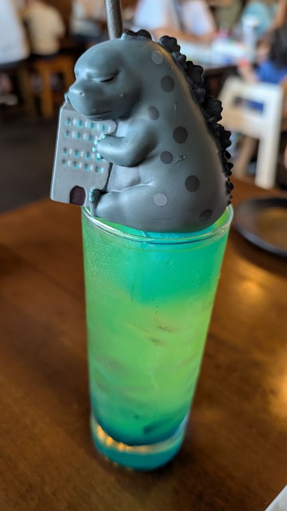 A small, smiling toy figure with a yellow hat sits on a wooden desk beside a glass of a green-colored drink garnished with lime slices and mint leaves.