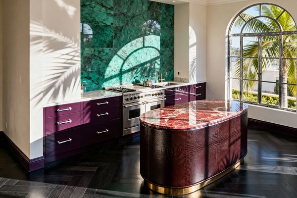 Sunlit kitchen with an arched steel-framed window casting palm shadows across cream walls and an ebony herringbone floor. A professional stainless range sits between plum lacquer base cabinets with chrome pulls and white marble tops, backed by a glossy emerald-green marble wall with a brass pot filler; an oval island wrapped in oxblood crocodile-textured leather with a red marble top rests on a brass plinth.