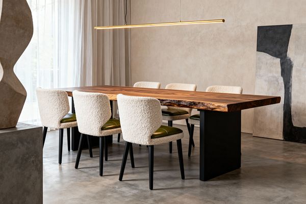A modern dining room features a long live-edge wood dining table on black steel pedestal legs, surrounded by six chairs with cream boucle backs, olive green leather seats, and black legs. A slim warm LED linear pendant hangs above, with floor-to-ceiling sheer curtains filtering daylight. The space has polished concrete floors, beige limewashed walls, a sculptural stone piece on a gray plinth, and a large leaning abstract black-and-white canvas.