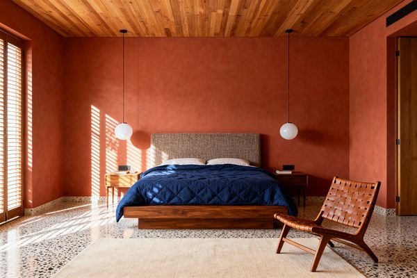 A mid-century modern bedroom features terracotta plaster walls, a tongue-and-groove wood ceiling, and a terrazzo floor. At center sits a low walnut platform bed with a tweed-upholstered headboard, navy quilt, and white pillows, flanked by walnut nightstands and globe pendant lights; sunlight filters through wooden louvered shutters, and a cognac woven-leather lounge chair and light rug soften the space.