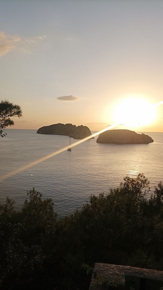 A serene sunset view over the ocean with two islands in the distance and a sailboat in the foreground.