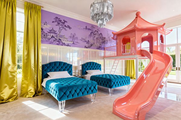 Bright kids’ bedroom with two teal, diamond‑tufted velvet twin beds on transparent acrylic legs, set against pearlized white wall panels and a lavender chinoiserie landscape mural. A coral pink, high‑gloss pagoda loft with slide and ladder anchors the right side, with chartreuse satin curtains, a cascading crystal‑chrome chandelier, tall windows and doors, and beige carpeting.