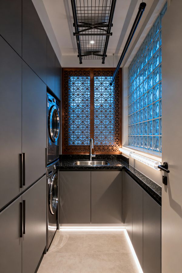 A narrow contemporary laundry room with matte gray cabinets and a U-shaped black speckled countertop. A stacked stainless washer and dryer are on the left; an undermount sink with a chrome gooseneck faucet sits below a window screened by geometric wood lattice. On the right, a glass-block wall casts cool blue light beside a tubular wall lamp, with ceiling-mounted black drying racks above and toe-kick LEDs illuminating light beige floor tile.