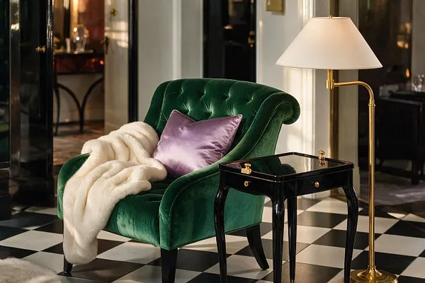 A Hollywood Regency seating vignette features a tufted emerald green velvet armchair with a lilac satin pillow and ivory faux-fur throw. Beside it sits a glossy black lacquer side table and a brass floor lamp with a white shade, all arranged over a black-and-white checkerboard floor in a polished, dramatic room.