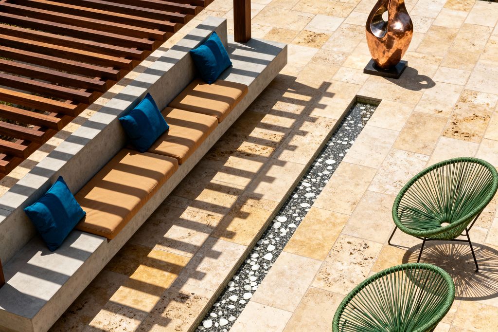 Sunlit patio with a long cast-concrete built-in bench under a slatted wood pergola that throws striped shadows across the seating. The bench has camel-colored seat cushions with deep teal throw pillows. The ground is paved in warm beige travertine tiles with a narrow linear channel filled with dark pebbles and white stones. Two round, green cord-woven lounge chairs sit on thin black metal legs, and a burnished copper sculpture stands on a small black plinth nearby.