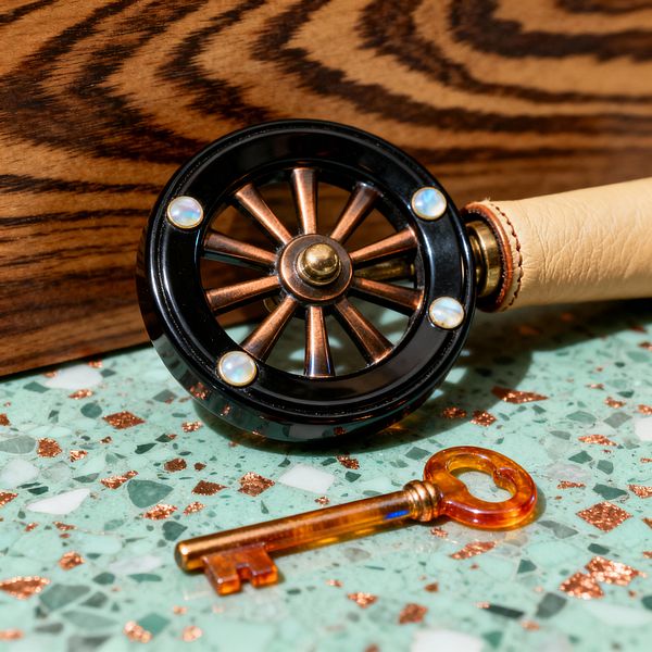 Close-up of an Art Deco-influenced ab roller resting on a mint green terrazzo surface with copper flecks. The wheel features a glossy black rim fastened with opalescent accents, copper spokes, and a brass hub, attached to a tan leather handle with visible saddle stitching. An amber key sits nearby, and a zebrawood panel with bold grain forms the backdrop, all lit warmly to emphasize metallic sheen and rich textures.