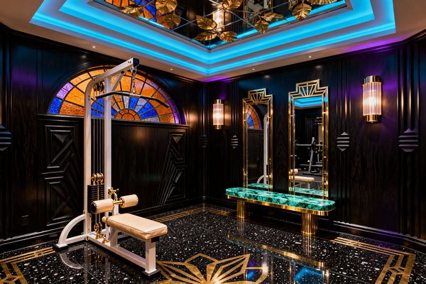 A luxurious Art Deco home gym with black paneled walls and geometric reliefs, brass-framed mirrors, and ribbed glass sconces, anchored by a semicircular stained-glass window in amber, cobalt, and violet above black terrazzo flooring with brass inlay and a starburst motif. A white weight machine with cream leather and brass pulleys faces an emerald malachite bench, while blue and purple cove lighting reflects off a mirrored ceiling soffit.