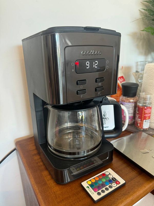 A coffee maker is displayed on a wooden surface, showing the time and a remote control nearby.