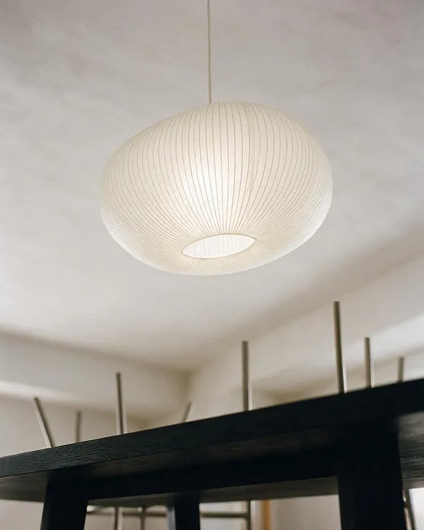 A modern interior ceiling light with a large oval paper lantern shade hanging from a thin cord. The soft ivory pendant has fine vertical ribbing and a circular opening on the bottom, set against a pale ceiling with a dark table edge visible below.