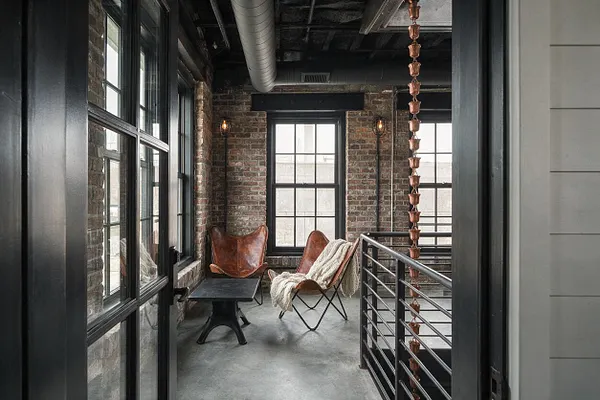 A narrow industrial front porch with exposed brick walls, black-framed windows, and a concrete floor. Leather butterfly chairs, a dark metal table, and a metal railing create a rugged, loft-like seating nook with a warm, understated feel.