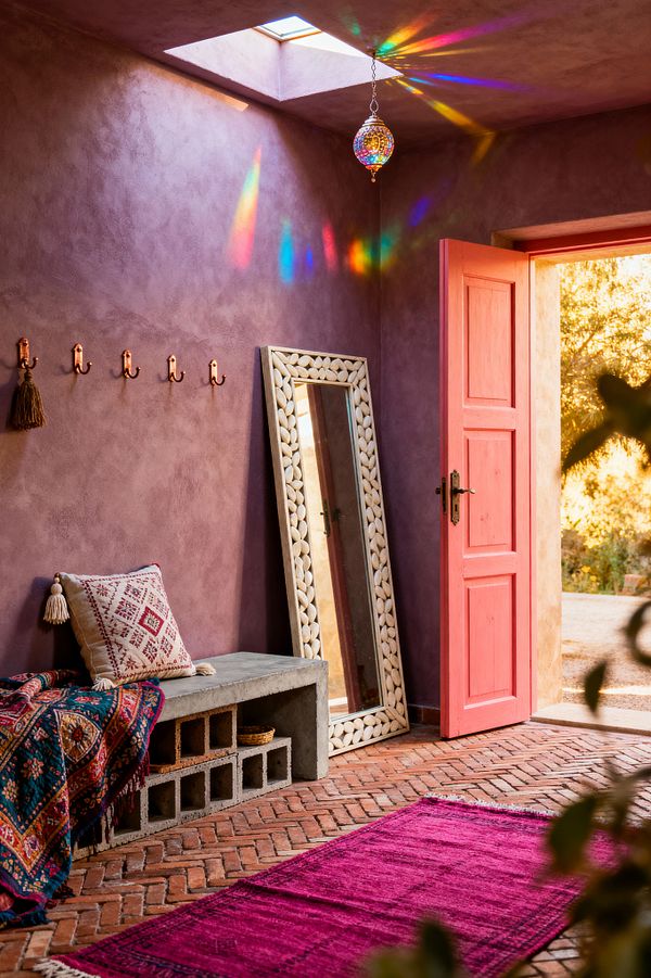 A bohemian mudroom with dusty mauve plaster walls features a coral-pink door open to golden light, a herringbone terracotta floor, and a concrete bench with cubbies draped in a patterned kilim and a tasseled cushion. A shell-inlaid full-length mirror leans against the wall beneath a skylight, while a multicolored glass pendant throws prismatic reflections; a magenta rug and copper hooks complete the warm, eclectic scene.