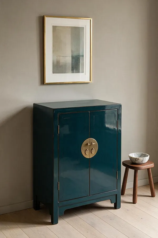 A glossy deep teal cabinet with two doors and a round brass medallion handle sits against a warm beige wall. Above it hangs a framed abstract artwork in a slim gold frame, and to the right a small wooden stool holds a white marble bowl on pale wood flooring.