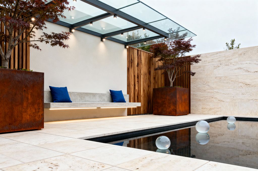 A sleek patio with a glass-roof canopy and small downlights shelters a long floating concrete bench lit by an LED strip, accented with two cobalt pillows. Light travertine tiles surround a narrow reflecting pool containing three translucent glass spheres. Large rusted Corten steel planters hold small maple trees, while a smooth plaster wall and vertical cedar slats create a clean, contemporary backdrop.