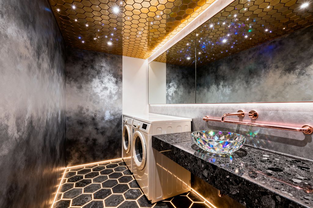 A dramatic laundry room features a gold hexagon-tiled ceiling dotted with star-like LEDs, mirrored by a long, backlit wall mirror. Smoky gray plaster walls and a glowing black hex tile floor frame a floating black terrazzo countertop with an iridescent glass vessel sink and rose-gold wall-mounted faucet; a white front-loading washer and dryer sit beneath bright task lighting, surrounded by warm ambient LED strips.