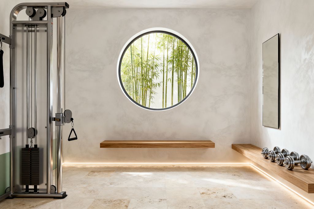 A minimalist home gym with warm white plaster walls features a large circular window centered on a view of bamboo. Creamy stone tiles cover the floor. Floating oak benches line two walls; one supports a row of chrome dumbbells. A slim rectangular mirror hangs on the right, and a stainless-steel cable machine stands on the left. Concealed LED strip lighting runs along the base and under the benches, casting a soft glow that emphasizes clean lines and open space.