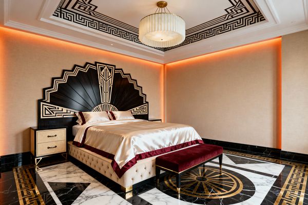 A luxurious Art Deco bedroom features a dramatic fan-shaped ebony headboard with gold geometric inlay behind a tufted cream bed dressed in champagne satin with burgundy trim. The space includes a burgundy velvet bench, cream nightstands with brass hardware, black-and-white marble flooring with gold Greek key borders and a sunburst medallion, a tray ceiling with black fretwork, warm cove lighting washing beige walls, and a pleated glass chandelier with a brass canopy.