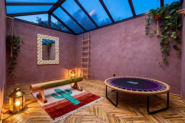 A bohemian home gym with mauve stucco walls sits under a sloped, blue-tinted skylight. A rope-edged mini trampoline, a natural log balance beam on a cactus-patterned rug, brass lanterns casting warm light, a pebble-framed wall mirror, a rope ladder in the corner, bamboo herringbone flooring, and trailing plants in terracotta hanging pots create an earthy, eclectic workout space.