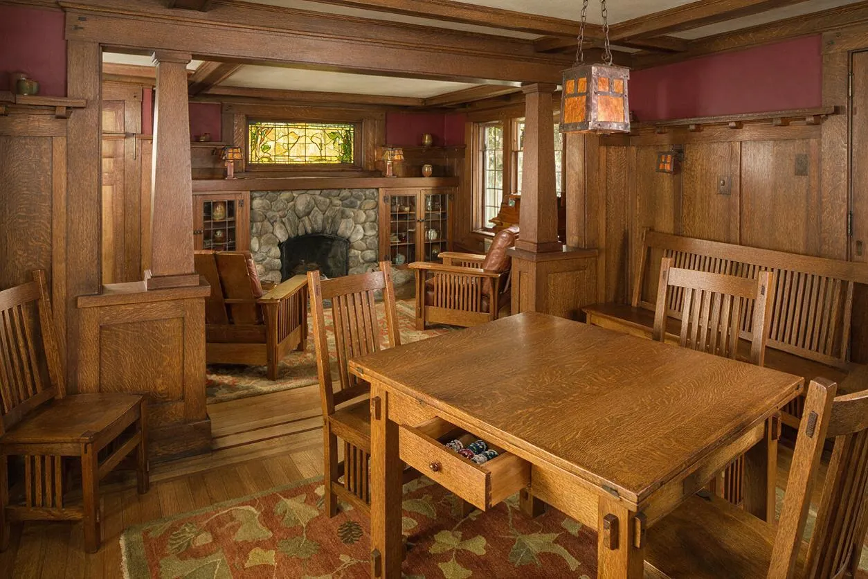 A Craftsman-style game room with extensive wood paneling, built-in benches, and a large wooden game table in the foreground. A stone fireplace, stained glass window, and amber pendant light add warmth and character.
