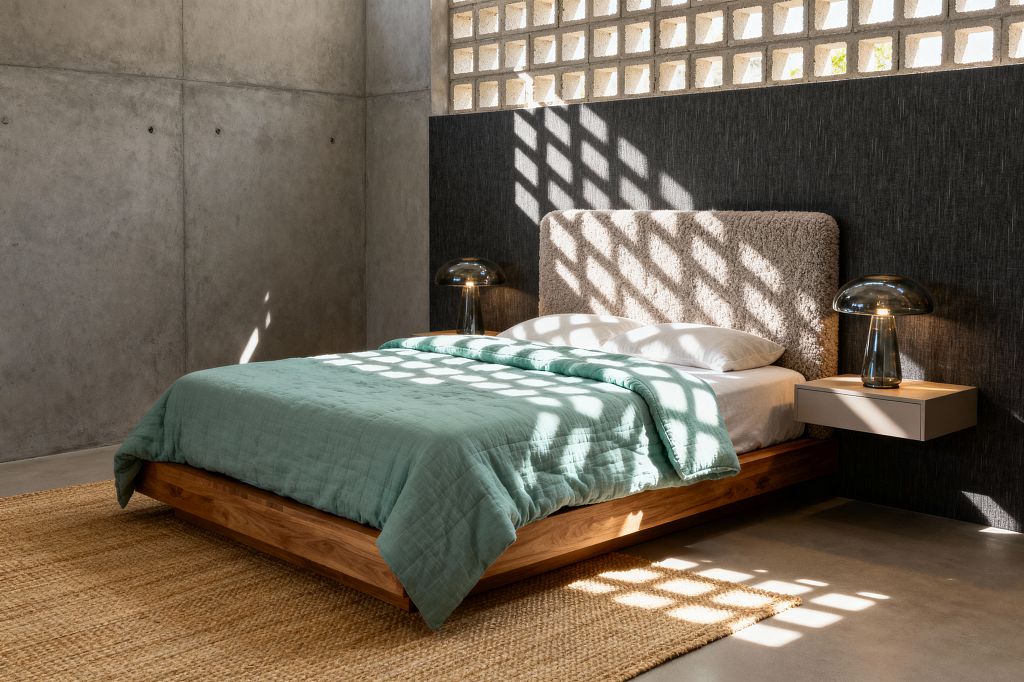 A minimalist mid-century bedroom with a low solid-wood platform bed and a light beige bouclé headboard set against a charcoal wall. Sunlight pours through a high breeze-block screen, casting a grid of shadows over seafoam bedding and white pillows, while matching smoked-glass mushroom lamps sit on bedside surfaces—one on a taupe floating nightstand—above a woven jute rug on polished concrete floors and raw concrete walls.