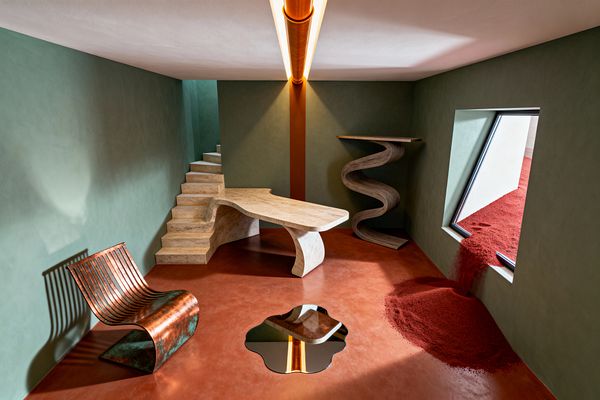 A surreal home office with sage-green plaster walls and a terracotta microcement floor. A sculptural travertine desk merges with a small staircase, next to an S-curved laminated wood shelf. An oxidized copper slatted lounge chair faces a puddle-shaped floor mirror. A warm tubular ceiling light aligns with a rust-toned vertical column, while a tilted black-framed window pours vivid red sand into the room.