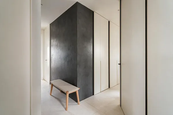 A minimalist entryway with smooth white walls, a tall charcoal-gray accent volume at the center, and light stone tile flooring. A small upholstered bench with pale wood legs sits in front, while flush white doors and slim black trim keep the space clean and calm.