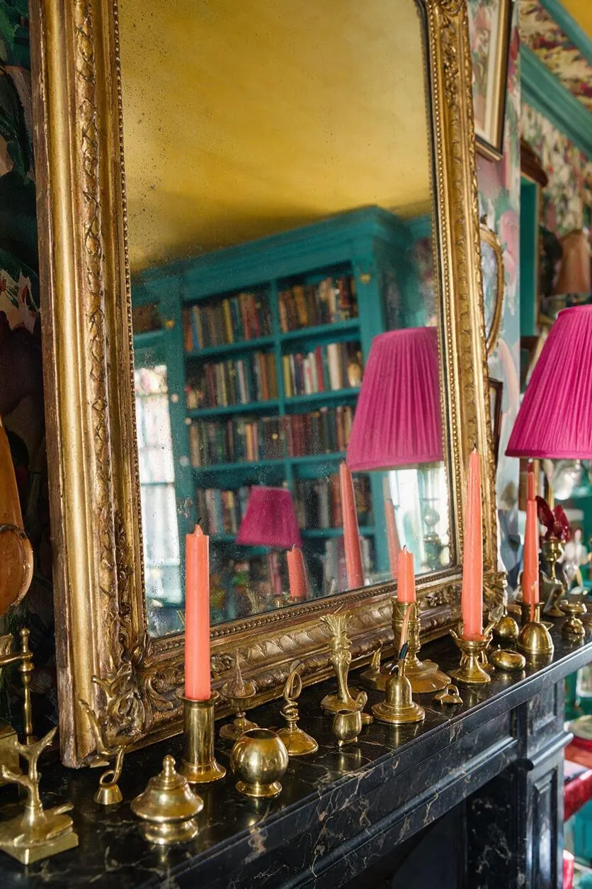 A large ornate gold-framed mirror rests above a black marble mantel covered with assorted brass decorative objects and coral candles. In the reflection, a turquoise bookcase, a yellow ceiling, and a bright pink lampshade reveal a richly layered maximalist room.