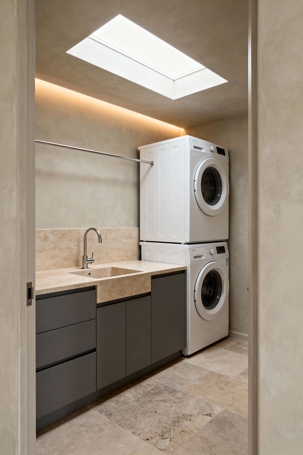 A compact minimalist laundry room shows a stacked white front-loading washer and dryer at the right, next to charcoal-gray, handleless base cabinets. The counter is honed beige travertine with an integrated square sink and a chrome gooseneck faucet, backed by a matching low backsplash. A slim stainless hanging rod spans the wall above. Large travertine floor tiles and smooth plaster walls set a warm neutral palette, while a ceiling skylight and concealed cove lighting provide even illumination.