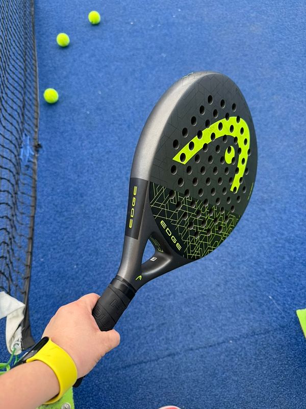 A hand holding a padel racket on a blue court with yellow balls in the background.