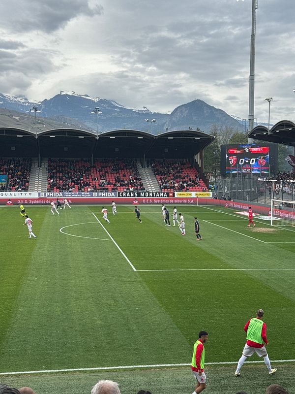 A soccer match is taking place with players from both teams on the field and a mountainous backdrop.