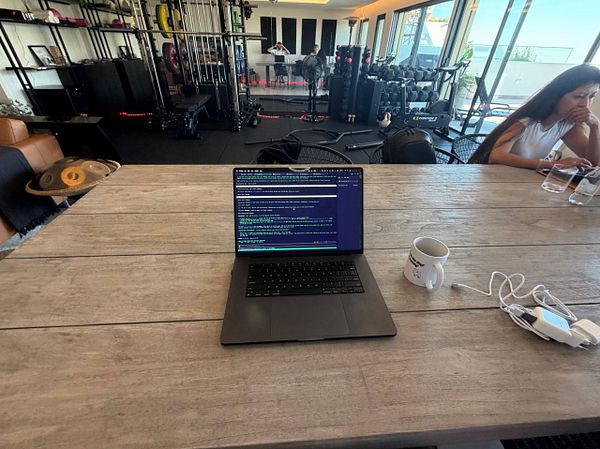 A coworking setup on a large wooden table in a room that also contains gym equipment, with a laptop displaying code, a coffee mug, and charging cables visible, and a person working on a laptop in the background.