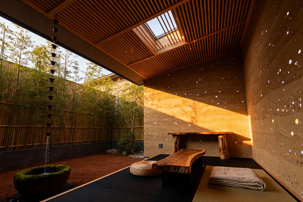 A minimalist indoor-outdoor living room features rammed-earth walls flecked with small reflective insets, a slatted wood ceiling with a central skylight, and a low live-edge wood bench beside a woven floor cushion and folded throw on tatami and black matting. A bamboo garden lines the open side, where a metal rain chain feeds a mossy stone basin, while warm sunset light washes the space with soft gold.