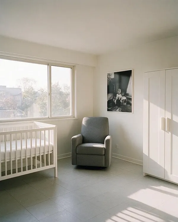 A bright minimalist nursery with white walls, a white crib near a large window, a gray upholstered armchair in the center, and a white wardrobe on the right. Soft natural light fills the room, creating a calm and uncluttered atmosphere.