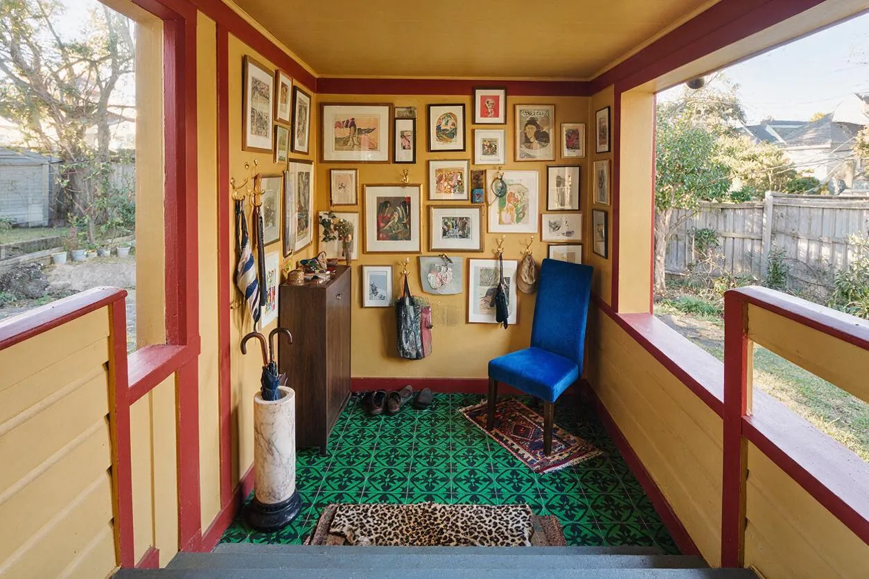 A small front porch styled in a maximalist way, with mustard yellow walls trimmed in deep red, a green patterned floor, and a dense gallery wall of framed artwork. A bright blue chair, umbrella stand, and layered doormats add color and personality, while the open sides reveal a fenced yard outside.