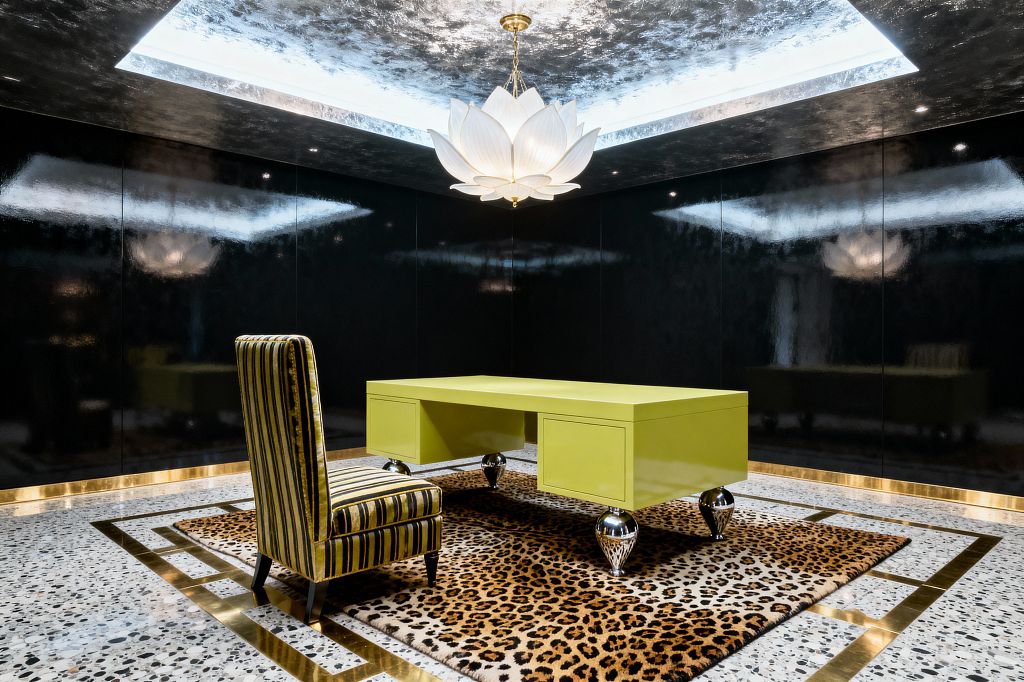 A dramatic Hollywood Regency home office features a chartreuse lacquered desk on polished chrome ball legs centered on a leopard-print rug framed by brass inlays on terrazzo flooring. A high-backed striped velvet side chair sits at the desk, while glossy black lacquer wall panels and gold base trim encircle the room. Overhead, a white lotus-petal chandelier hangs within a silver-leaf coffer with concealed lighting, creating reflective highlights that emphasize the room’s bold contrasts and glamorous materials.