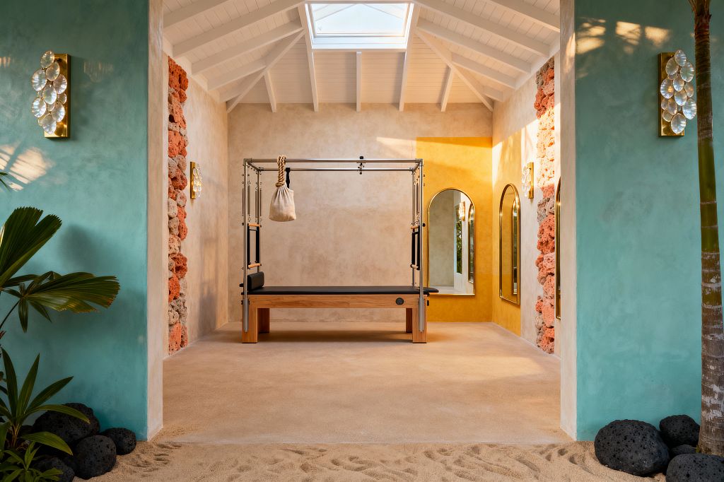 A bright tropical home gym features a Pilates Cadillac with an oak base and brushed metal frame under a white vaulted ceiling with a skylight. Limewashed aqua side walls reveal bands of coral stone and lead to a mustard-ochre alcove with arched brass-framed mirrors; capiz-shell sconces glow on the walls. The sandy microcement floor transitions to real sand with black lava rocks and a palm, creating an indoor-outdoor, beach-inspired feel.