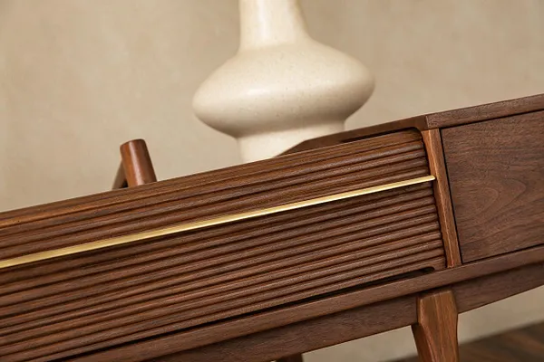 A close-up of a mid-century modern wooden cabinet or credenza with richly grained walnut, horizontal reeded drawer fronts, and a slim brass accent line. Tapered wooden legs and a soft sculptural vase in the background reinforce the warm, streamlined style.